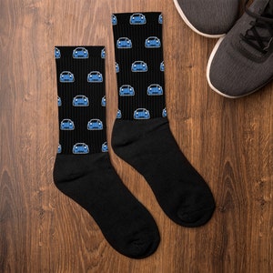 Könnte beinhalten: Schwarze Socken mit einem sich wiederholenden Muster aus blauen Autoillustrationen. Die Socken werden auf einer Holzoberfläche neben einem Paar grauer Turnschuhe gezeigt. Das Autodesign hat einen blauen Körper und weiße Akzente.