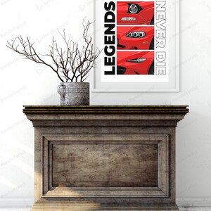 May include: A framed print featuring three red car front-end illustrations. The print has the words "LEGENDS" and "NEVER DIE" in black and white. The print is displayed on a dark wooden cabinet, with a decorative vase holding branches.