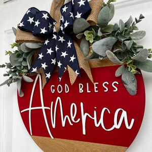 May include: A red and brown wooden round door hanger with the text "God Bless America" in white lettering. The hanger is decorated with a blue and white star patterned bow and green foliage.