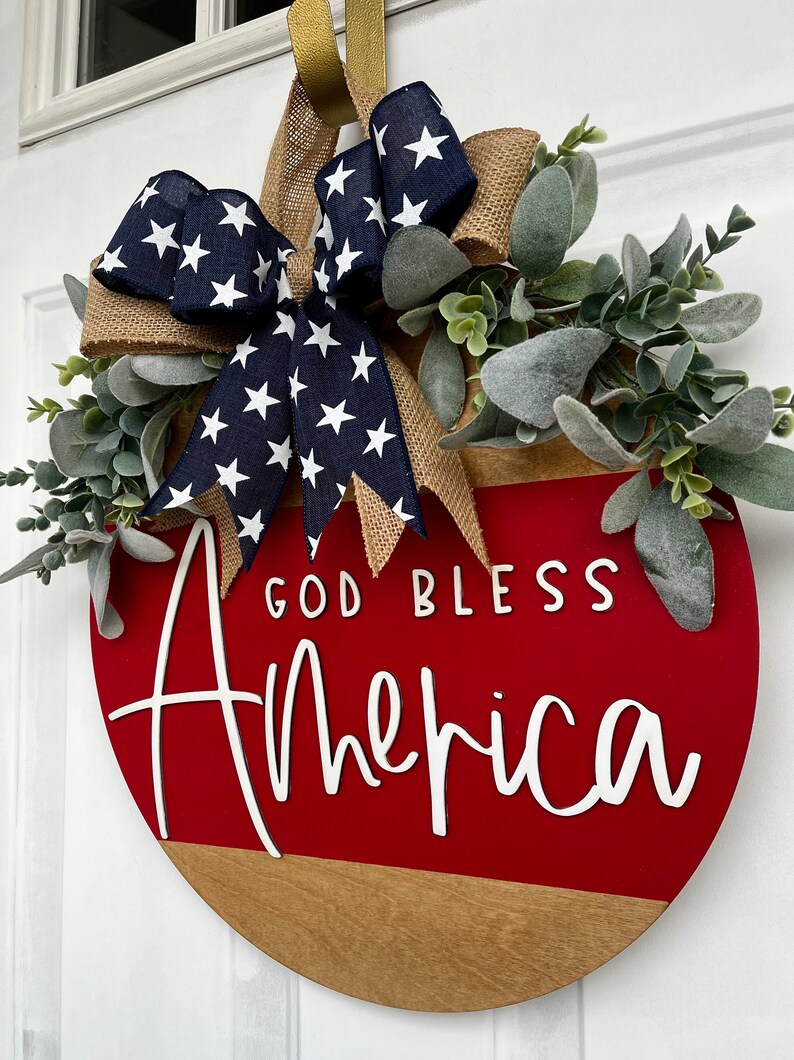 May include: A red and brown wooden round door hanger with the text "God Bless America" in white lettering. The hanger is decorated with a blue and white star patterned bow and green foliage.