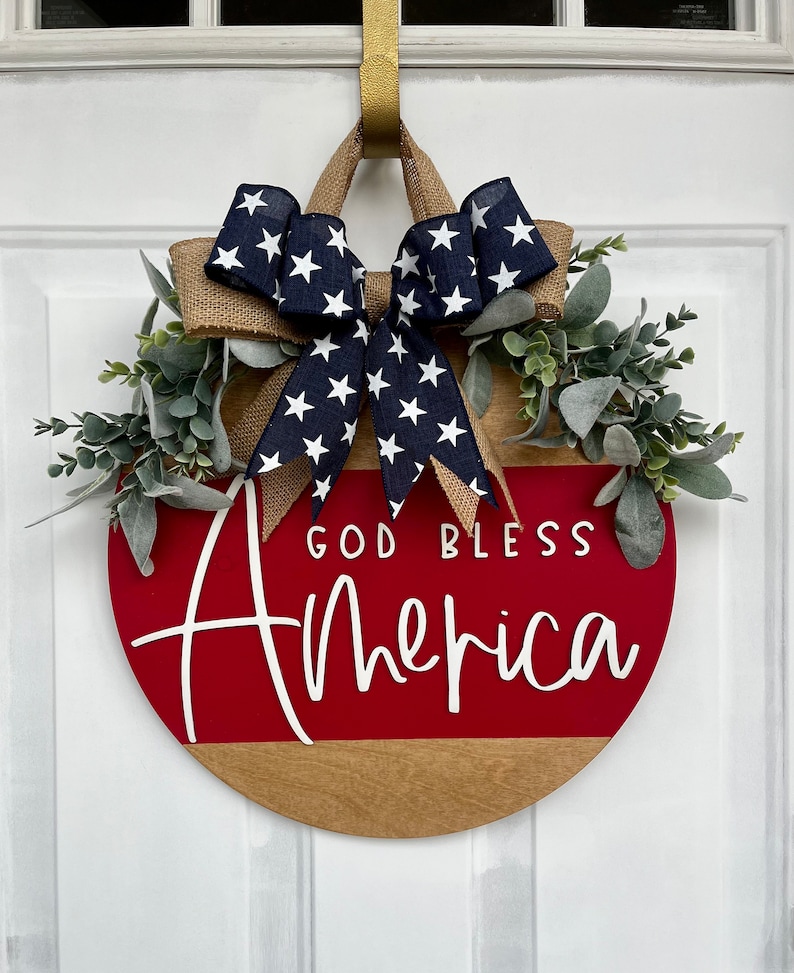May include: A red and brown wooden door hanger with the text "God Bless America" in white lettering. The hanger is decorated with a blue and white star patterned bow and a green leafy garland.