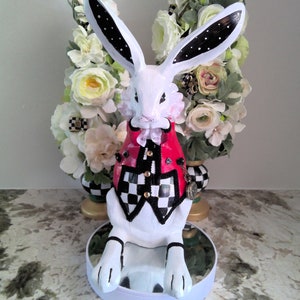 Könnte beinhalten: Weiße Keramik-Hasenfigur, die eine rot-schwarz karierte Weste mit goldenen Akzenten trägt. Der Hase sitzt auf einem weißen Sockel mit einer spiegelnden Oberfläche.