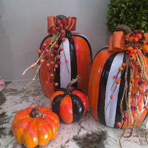 May include: Three decorative pumpkins with black and white stripes, orange and black stripes, and orange and black stripes. The pumpkins are decorated with gold glitter, red and orange berries, and orange and black bows.