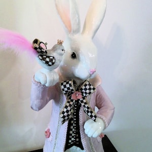 Può includere: Una statuetta di coniglio bianco che indossa una giacca rosa con un fiocco a scacchi bianco e nero e una piccola statuetta di uccello posata sulla sua mano. Il coniglio ha le orecchie rosa e un naso rosa.