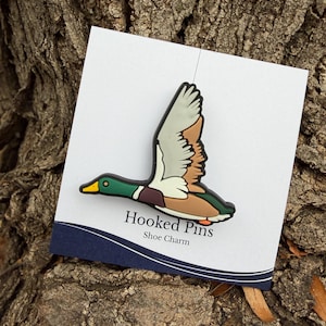 May include: A shoe charm featuring a flying duck with green, brown, and white feathers, a yellow beak, and orange feet. The charm is attached to a white card with the text "Hooked Pins Shoe Charm" in black.