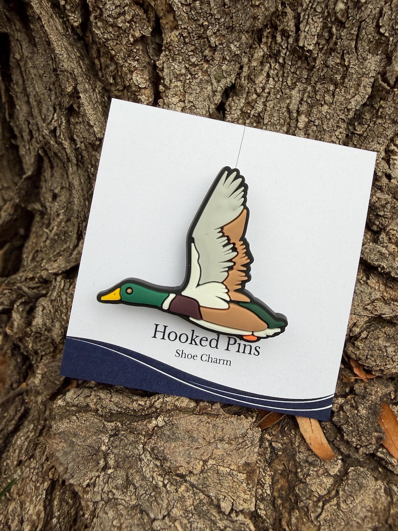 Op de afbeelding: Een schoenbedel met een vliegende eend met groene, bruine en witte veren, een gele snavel en oranje poten. De bedel is bevestigd aan een witte kaart met de tekst "Hooked Pins Shoe Charm".