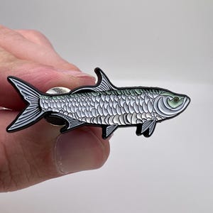 May include: A detailed enamel pin in the shape of a fish. The pin features a silver body with black outlines, a black tail, and a green eye. The fish pin is a unique accessory for jackets, bags, or hats.