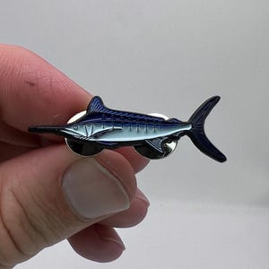 Könnte beinhalten: Ein blau-weißes Marlin-Anstecknadel mit schwarzer Umrandung. Die Anstecknadel zeigt ein detailliertes Design des Fisches, einschließlich seines langen Schnabels und der markanten Rückenflosse. Die Anstecknadel ist aus Metall und hat ein glänzendes Finish.