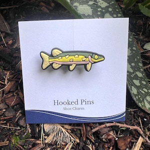 Könnte beinhalten: Ein schuhförmiger Anhänger in Form eines Fisches mit grünem Rücken, gelbem Bauch und schwarzer Umrandung. Der Anhänger befindet sich auf einer weißen Karte mit dem Text "Hooked Pins Shoe Charm".