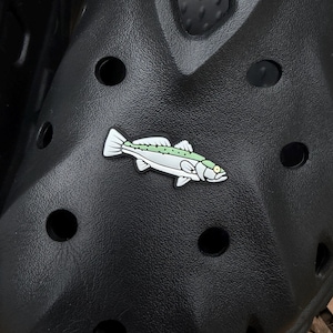 May include: A white, green, and black fish-shaped shoe charm. The charm is affixed to a black shoe with multiple holes. The fish has a detailed design, including fins, scales, and an eye. The shoe has a textured surface.