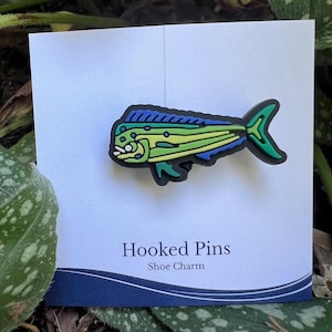 May include: A colorful shoe charm featuring a mahi-mahi fish design. The fish is primarily green and blue with yellow accents, set against a white card with the text "Hooked Pins Shoe Charm".