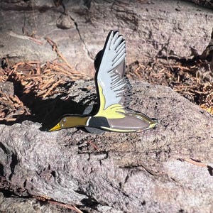 May include: A detailed enamel pin depicting a duck in flight. The pin features a yellow beak, a green head, and a brown body with black and yellow accents. The wings are white and gray. The pin is set against a natural, textured background.