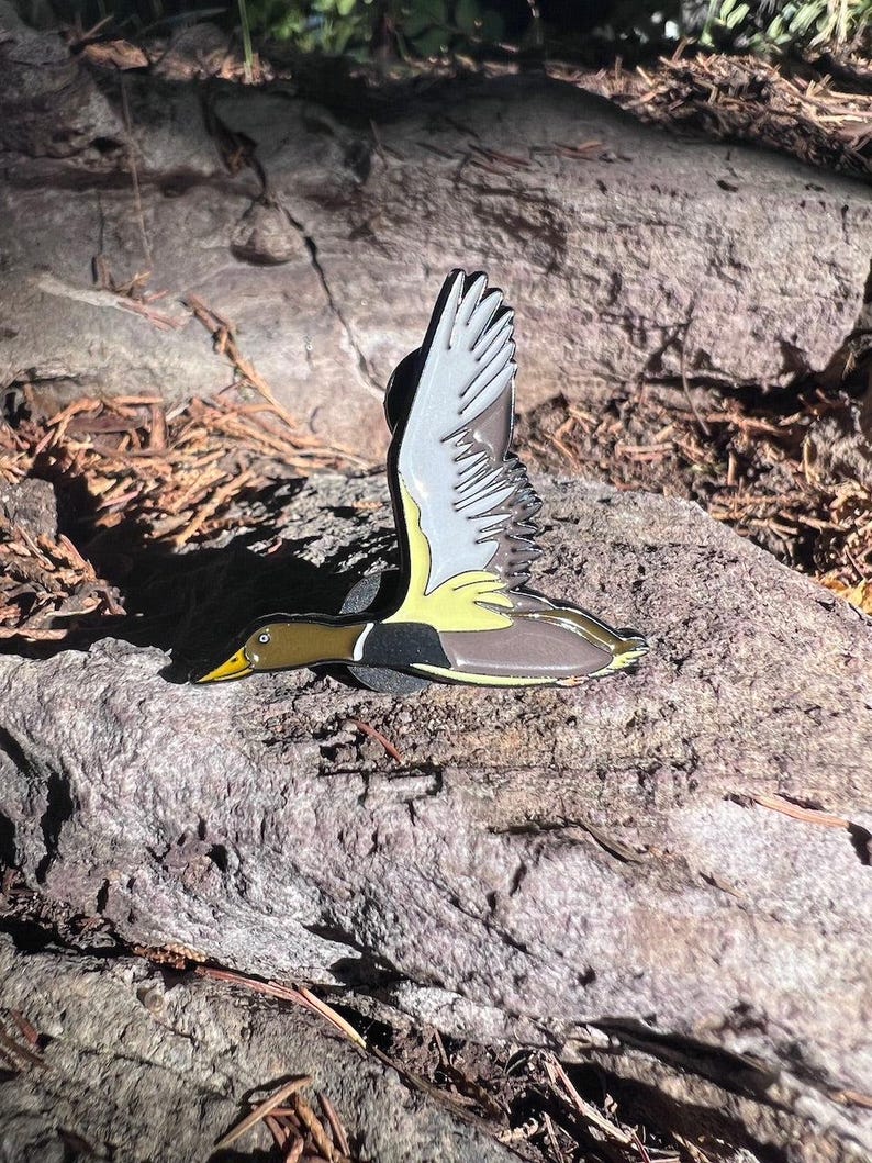 May include: A detailed enamel pin depicting a duck in flight. The pin features a yellow beak, a green head, and a brown body with black and yellow accents. The wings are white and gray. The pin is set against a natural, textured background.