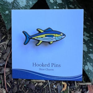 May include: A colorful fish-shaped shoe charm with blue, yellow, and white accents. The charm is on a white card with the text "Hooked Pins Shoe Charm". The background includes greenery and wood chips.