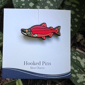 May include: A shoe charm shaped like a red and black fish with a white belly and green eye. The charm is on a white card with the text "Hooked Pins Shoe Charm". The background is green foliage.