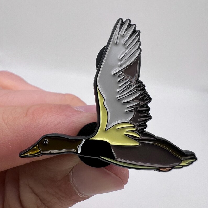 May include: A detailed enamel pin depicting a duck in flight. The pin features a brown body, yellow accents, and gray and white wings. The duck is shown mid-flight, with its wings spread. The pin has a black backing.