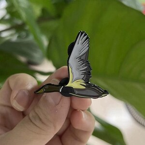 May include: A detailed enamel pin depicting a duck in flight. The pin features a black body, gray and white wings, and yellow accents. The duck pin is a nature-themed accessory.