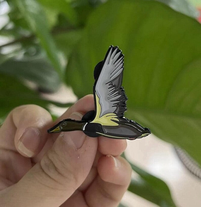 May include: A detailed enamel pin depicting a duck in flight. The pin features a black body, gray and white wings, and yellow accents. The duck pin is a nature-themed accessory.