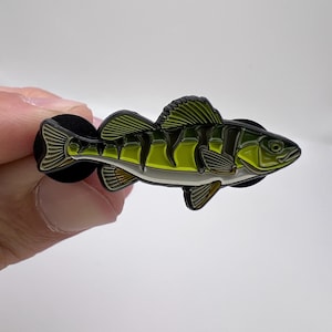 以下が含まれることがあります： 黄色いパーチ魚の形をした、詳細なエナメルピン。 ピンは、魚の鱗とひれを示す、黒いアクセントが付いた緑と黄色の配色です。 ピンは白い背景に対して手で保持されています。