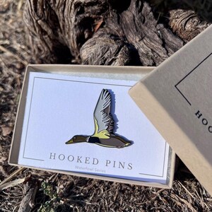May include: A waterfowl pin in a gift box. The pin features a flying duck with gray, brown, and yellow details. The box has the text "HOOKED PINS Waterfowl Series" on a white card.