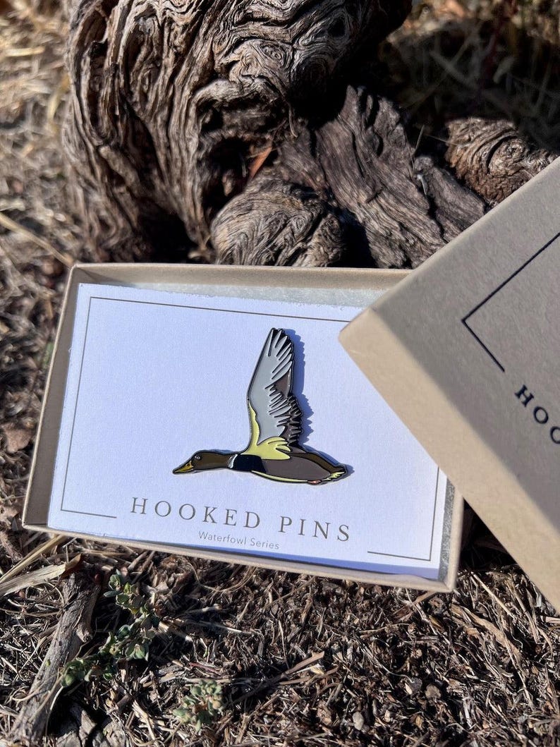 May include: A waterfowl pin in a gift box. The pin features a flying duck with gray, brown, and yellow details. The box has the text "HOOKED PINS Waterfowl Series" on a white card.