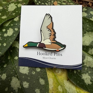 Op de afbeelding: Een schoenbedel met een vliegende wilde eend. De eend heeft een groene kop, een bruin lichaam en witte en bruine vleugels. De bedel is bevestigd aan een witte kaart met de tekst "Hooked Pins Shoe Charm" in het zwart.