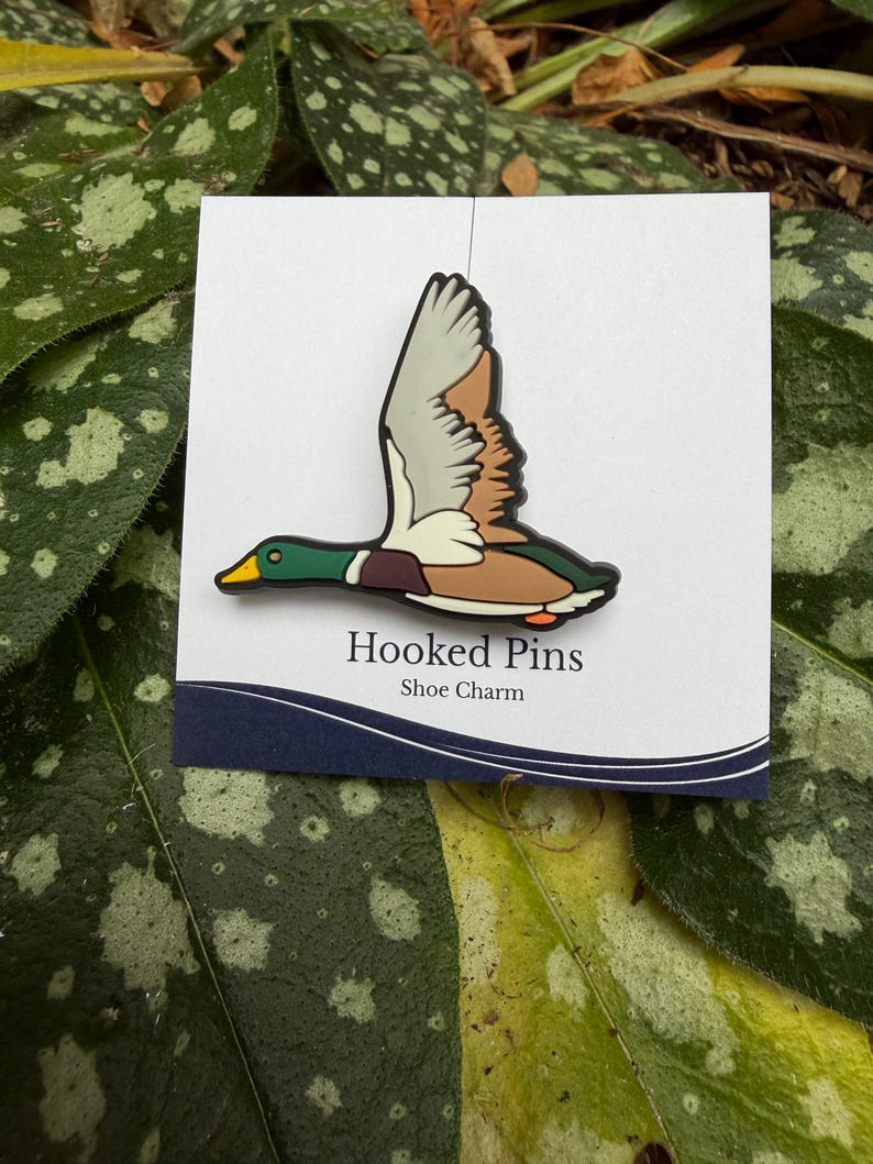 Op de afbeelding: Een schoenbedel met een vliegende wilde eend. De eend heeft een groene kop, een bruin lichaam en witte en bruine vleugels. De bedel is bevestigd aan een witte kaart met de tekst "Hooked Pins Shoe Charm" in het zwart.