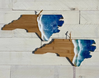 May include: Two wooden chopping boards shaped like the state of North Carolina. The boards are decorated with blue and white resin to resemble ocean waves.