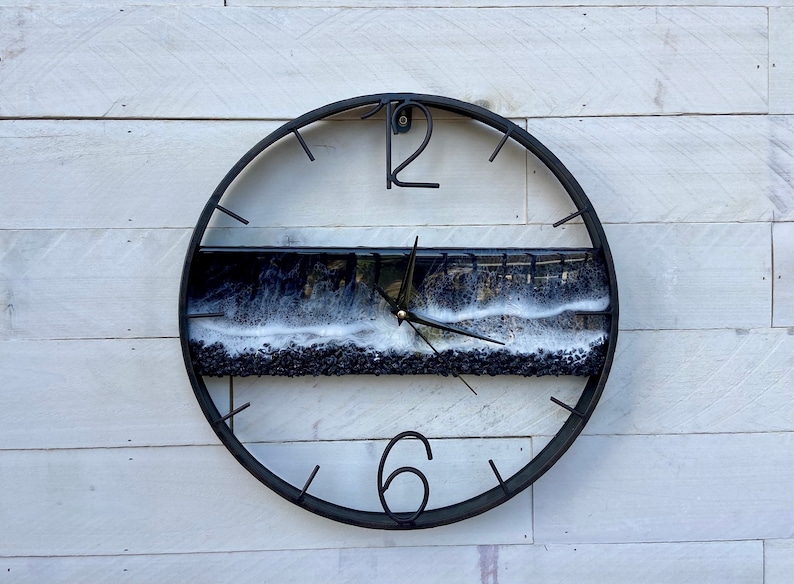 May include: A black circular clock with a black and white resin insert featuring a wave design. The clock face has black Roman numeral markings for the hours.