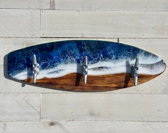 May include: A wooden surfboard-shaped wall hook with a blue and white resin ocean wave design. The hook has three silver metal cleat hooks.