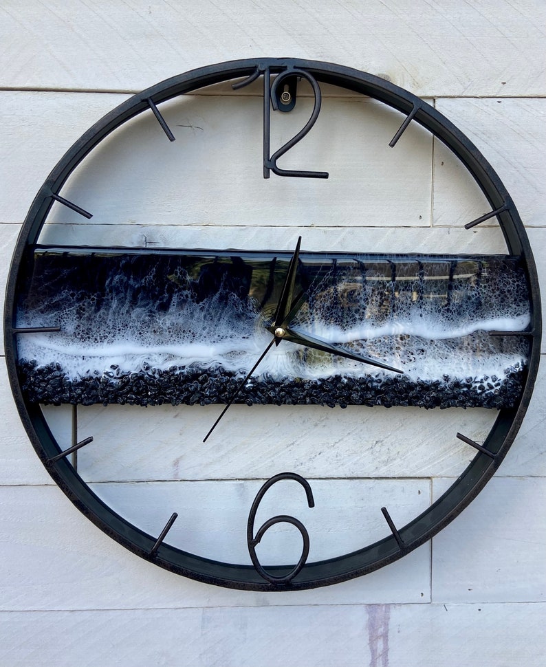 May include: A black metal clock with a white and black resin wave design. The clock face has black Roman numeral markings and black hands.