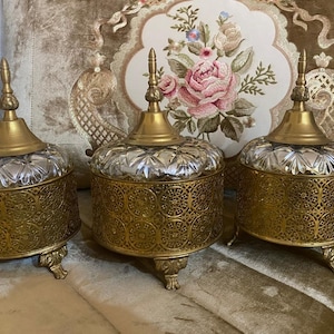May include: Three decorative antique jars with ornate gold filigree bases and lids. The jars have clear, faceted glass tops and are set against a floral embroidered background. The jars have a vintage aesthetic.