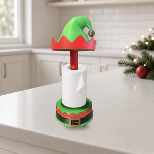 May include: A toilet paper holder featuring a red central pole, a green and red elf hat topper with a small bell, and a green base with a black belt and gold buckle design. The holder supports a single roll of white toilet paper.
