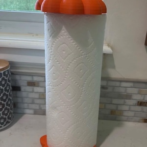 May include: A paper towel holder featuring a bright orange pumpkin-shaped base and a matching pumpkin-shaped top cap with a brown stem. The holder is designed to hold a standard roll of white paper towels with a textured diamond pattern.