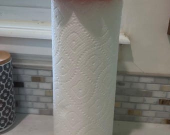 May include: A paper towel holder featuring a bright orange pumpkin-shaped base and a matching pumpkin-shaped top cap with a brown stem. The holder is designed to hold a standard roll of white paper towels with a textured diamond pattern.