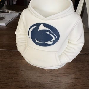 May include: A white 3D-printed container shaped like a miniature hoodie. The front features a dark blue oval emblem with a stylized white mountain lion silhouette. The design includes a hood, long sleeves, and a front kangaroo pocket.