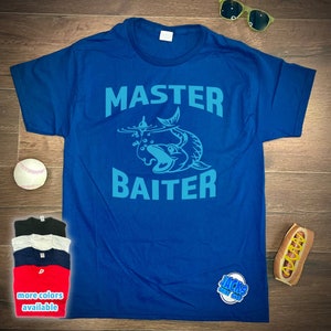 May include: A blue t-shirt with the text "MASTER BAITER" in a light blue color. A cartoon fish is biting a fishing lure. The text "JACKS SHIRT SHOP" is in the bottom right corner.