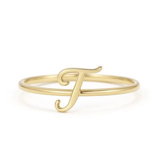 May include: A delicate gold ring featuring the cursive letter "F". The ring has a thin band and the letter is the focal point, with a smooth, polished finish. The ring is set against a white background.