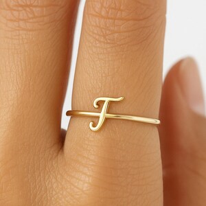 Pode incluir: Um delicado anel dourado com a letra cursiva "F". O anel é fino e minimalista, com a letra como elemento principal do design. O anel é mostrado num dedo, realçando a sua elegância e simplicidade.