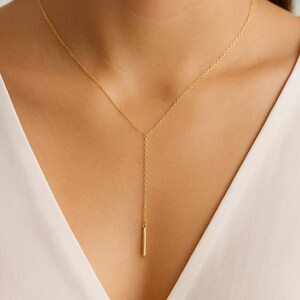 Puede incluir: Un delicado collar dorado con un colgante de barra vertical. El collar se muestra sobre un fondo blanco, resaltando la simple elegancia de la joya. La cadena es fina y el colgante de barra cuelga con gracia.