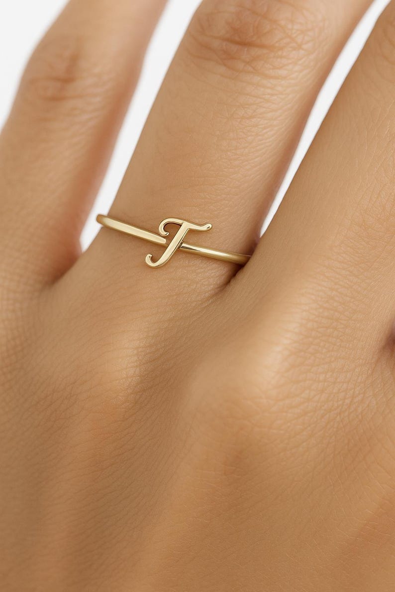 May include: A gold ring with the letter "F" in a cursive style. The ring is slender and minimalist, with the letter as the primary design. The ring is displayed on a finger.
