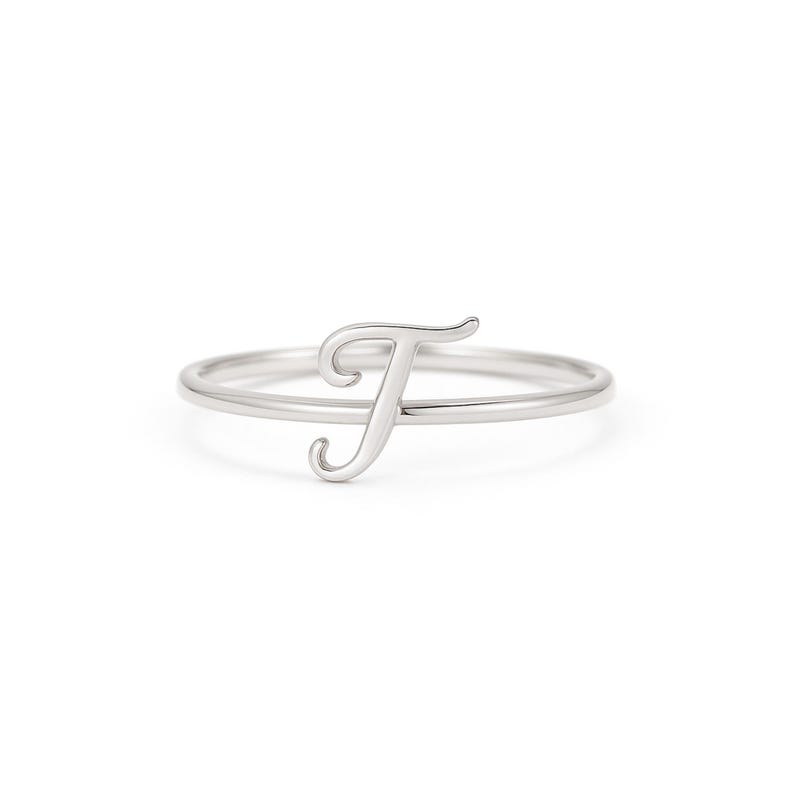 May include: A silver ring with the cursive letter "T". The ring has a slender band and the letter is the main feature, with a graceful, flowing design. The ring is set against a white background.