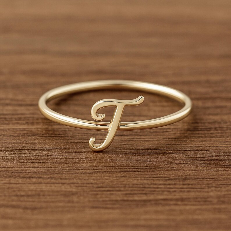 May include: A delicate gold ring featuring the letter "T" in a cursive font. The ring has a thin band and the letter is the focal point, creating a minimalist and elegant design. The ring is shown on a wooden surface.