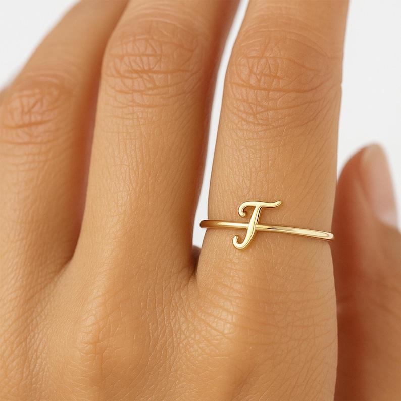 May include: A delicate gold ring displaying the cursive letter "F". The ring has a thin, minimalist design, with the letter as the main feature. The ring is shown on a finger, emphasising its elegant simplicity.
