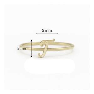 May include: A gold ring with the letter "T". The letter and band are each 5 mm wide. The ring is displayed against a white background.