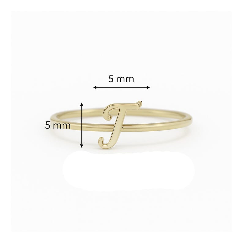 May include: A gold ring with the letter "T". The letter and band are each 5 mm wide. The ring is displayed against a white background.