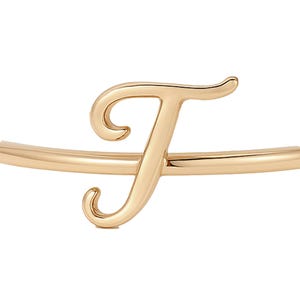 May include: A gold-coloured bangle bracelet featuring the letter "F" in a cursive font. The letter is the focal point, with a smooth, polished finish. The bracelet is a delicate piece of jewellery.