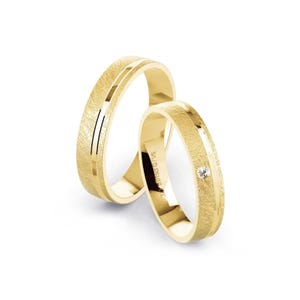 May include: Two gold wedding bands. The bands have a textured surface with a smooth, central groove. One ring features a small, clear gemstone. The rings are set against a white background.