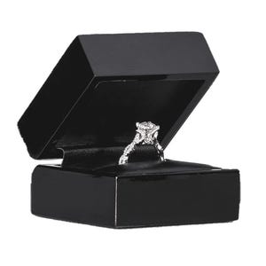 May include: A diamond engagement ring presented in an open black jewellery box. The ring has a large, round-cut diamond on a detailed, silver band. The box is glossy with a black interior.