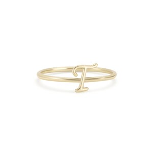 May include: A gold-coloured ring with the letter "T" in a delicate, cursive font. The ring has a thin band and the letter is raised, creating a minimalist and elegant design. The ring is likely made of gold or a gold-coloured metal.